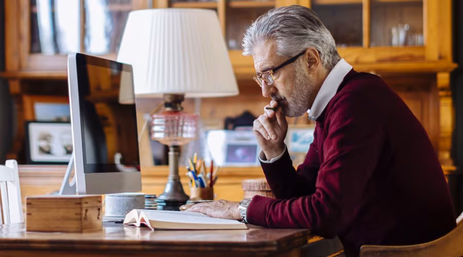 An older gentleman in a burgundy sweater sits at a wooden desk, intently reading a book in a warmly furnished office.
