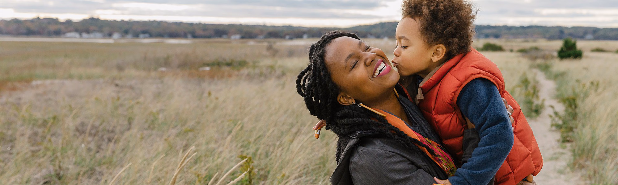 A young woman laughs and leans back in a grassy field as the child in her arms leans in to kiss her cheek.